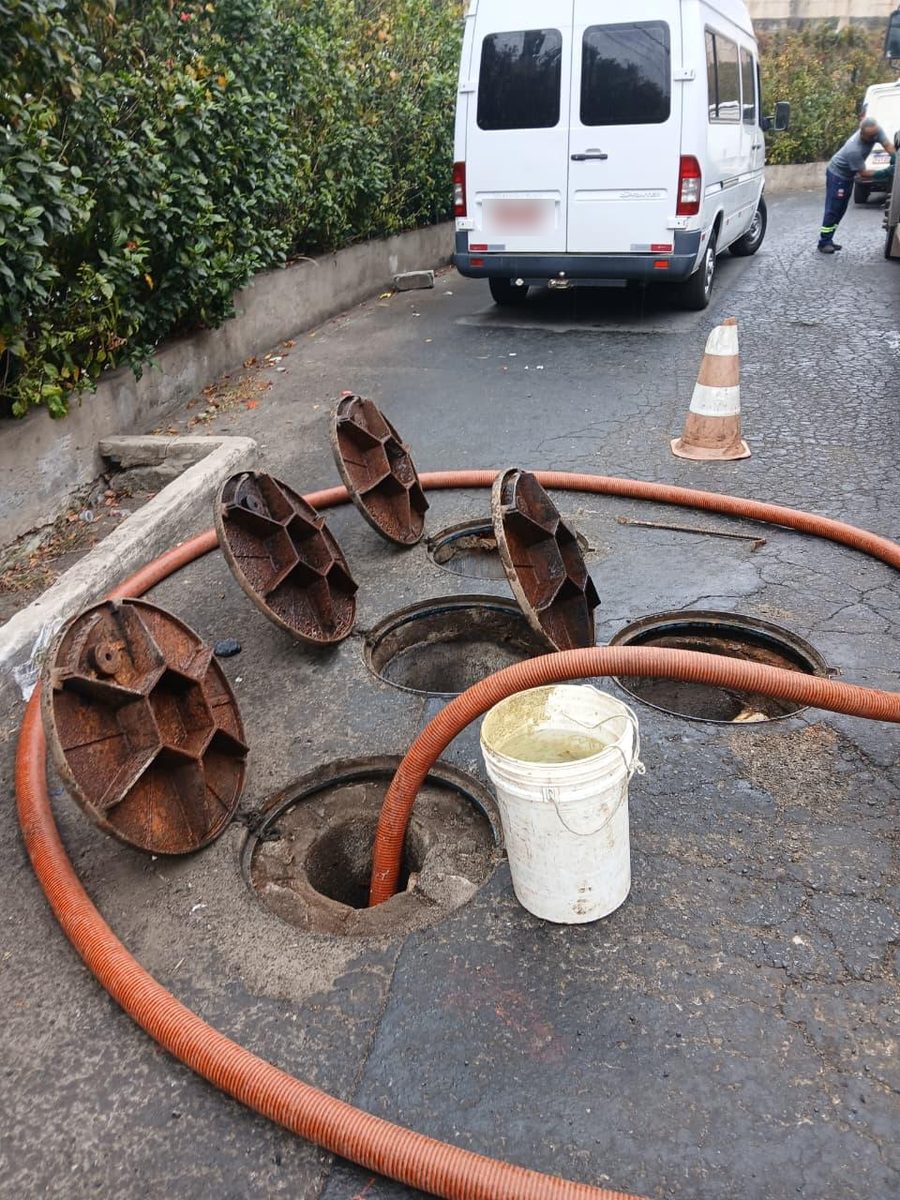 Tampas de caixas removidas e mangueira de sucção inserida em uma das aberturas, com um funcionário ao fundo